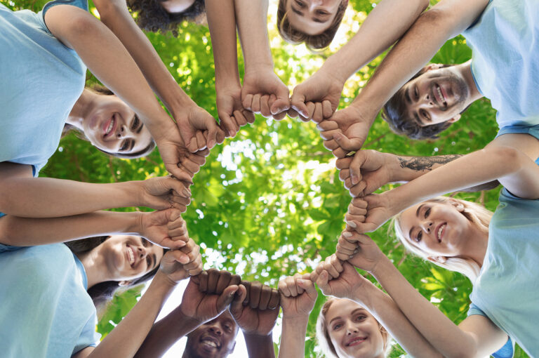 Gruppo di persona con magliette azzurre, in un cerchio, al centro del cerchio si ha come sfondo gli alberi verdi. La visione è dal basso.